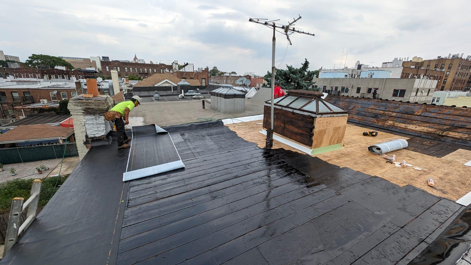 Two crew members installing a low-slope roof membrane over rebuilt decking on a Brooklyn home