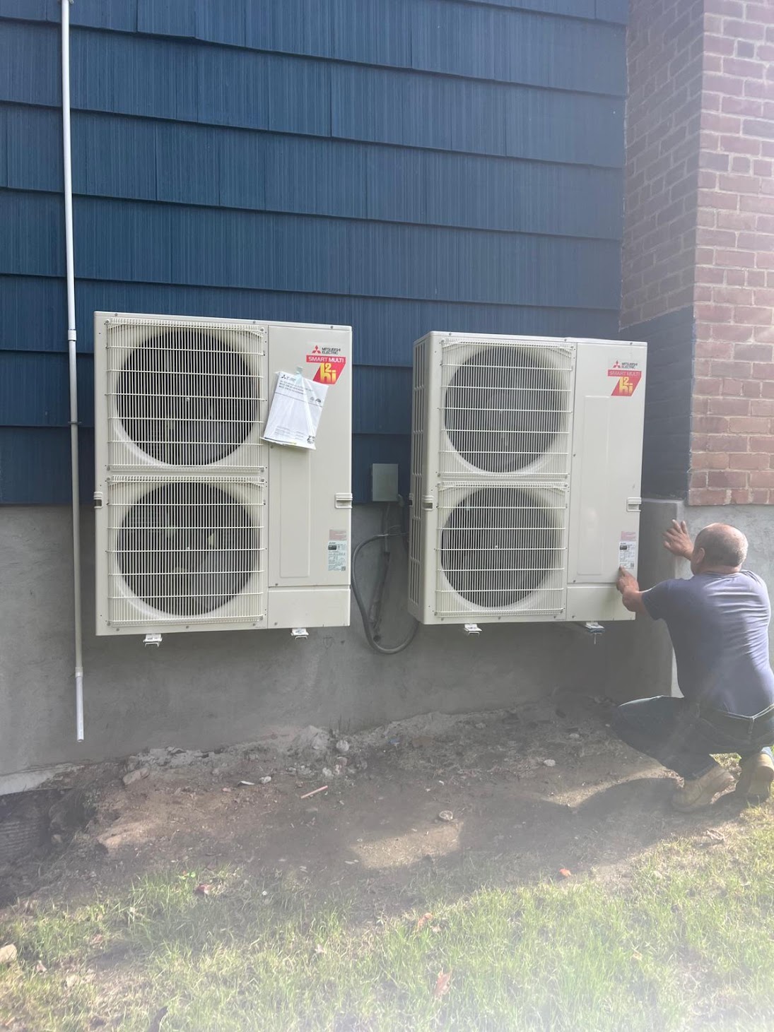 Technician working beside two exterior heat-pump condensers on a Kellets project