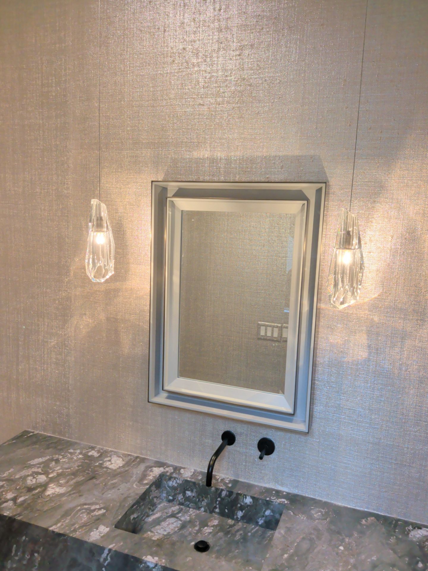 Finished Scarsdale powder room with textured metallic wallcovering, stone vanity, mirror and decorative sconces